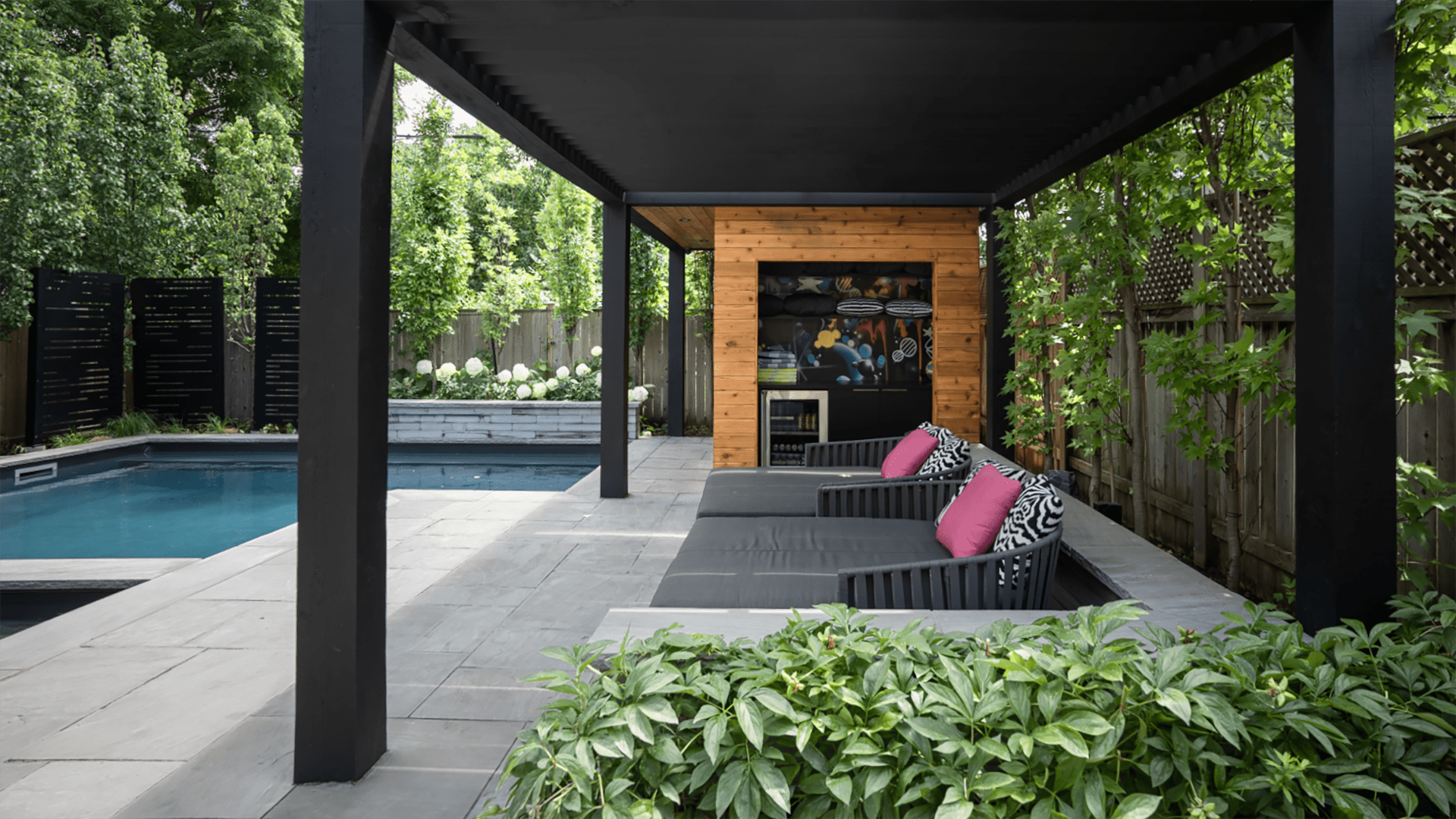 A modern backyard with a pool, pergola, and lounge chairs featuring pink cushions, surrounded by lush greenery and wooden fencing.